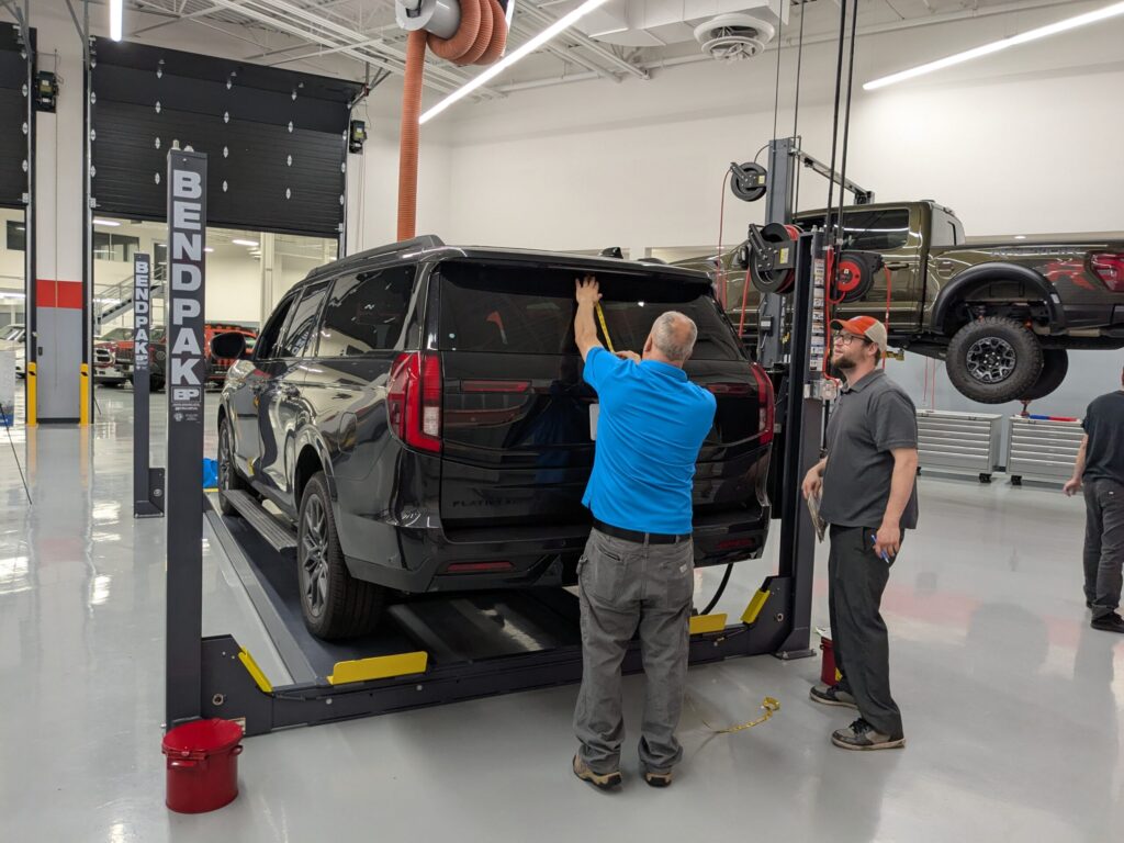 Technician measuring the rear liftgate of a black SUV on a vehicle lift, while another team member observes and takes notes inside the SEMA Garage.