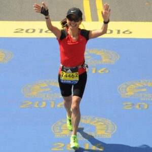 Victoria Repice smiling with arms raised as she crosses the finish line at the 2016 Boston Marathon, wearing a red running shirt, black shorts, and race bib 24462.