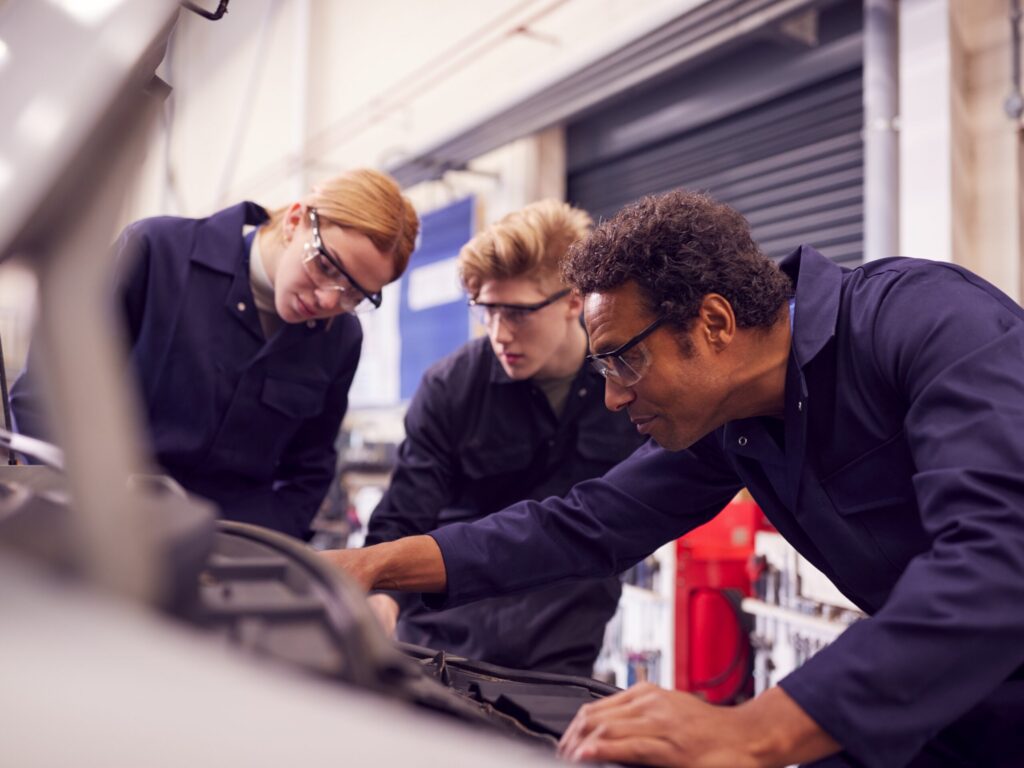 An experienced automotive instructor demonstrates engine diagnostics to two students in a professional repair workshop. All three wear navy coveralls and safety glasses while examining the open hood of a vehicle, highlighting hands-on training in automotive repair and technical education.