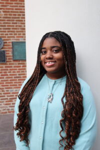 Damikka Willis, Marketing Intern at MOTOR, smiling in a professional headshot with long braids, light blue sweater, and silver cat necklace, posed against a brick wall and white column.