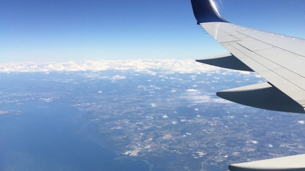 View from airplane window showing wing over landscape and coastline with scattered clouds below.