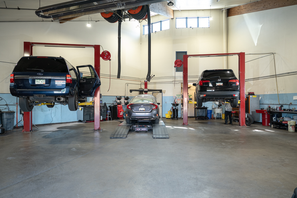 Three service bays inside the Autovisions repair facility, with two vehicles on lifts and one centered on the ground. The shop is clean, well-lit, and actively in use.
