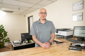 Nathan Bryant standing behind the front counter of his shop, hands folded, with a welcoming expression. Office equipment and service certifications are visible in the background.