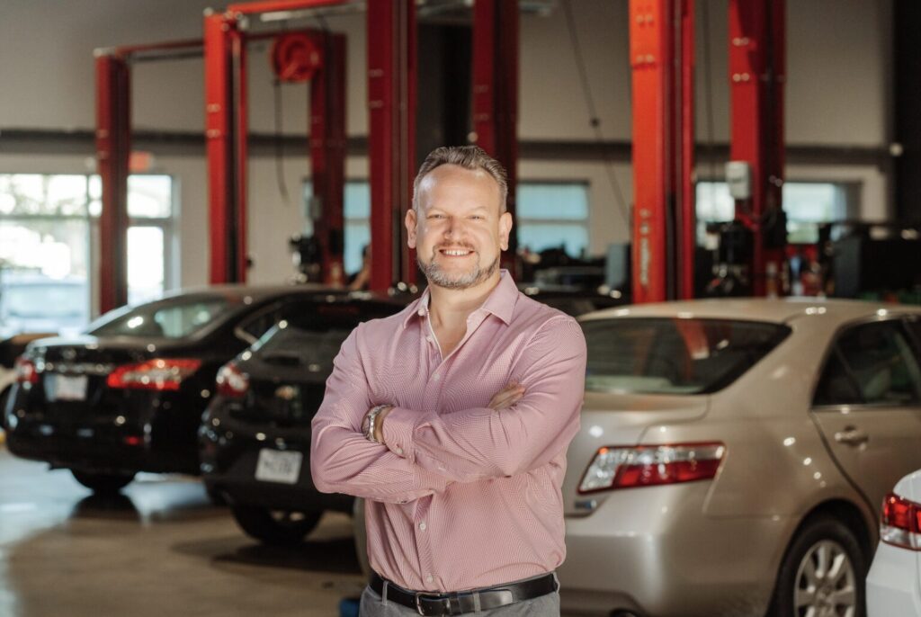 Jack Getz smiling with arms crossed inside Getz Automotive repair shop, standing in front of cars and red service lifts.