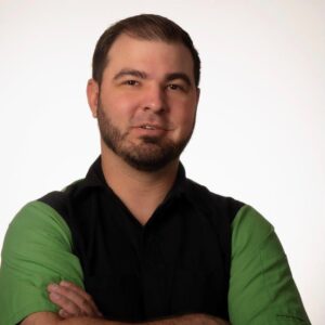 Portrait of a man with short dark hair and a neatly trimmed beard, wearing a black shirt with green sleeves, standing against a plain white background with arms crossed and a confident expression.