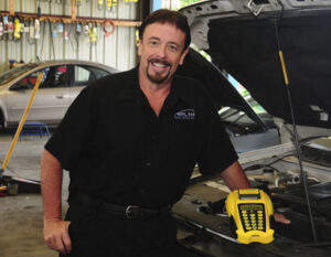Smiling male automotive technician standing next to an open vehicle hood in a repair shop, wearing a black shop shirt embroidered with a logo and holding a yellow diagnostic scan tool. Tools and a silver car are visible in the background.