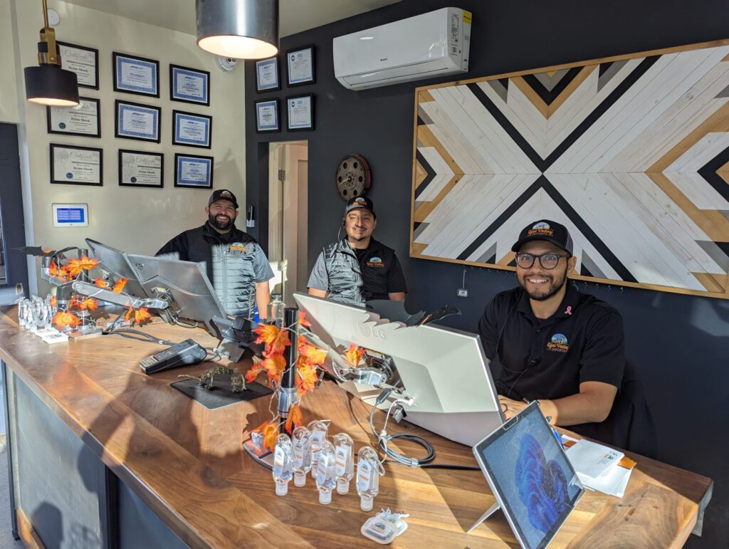 Service advisors at LBM Automotive smiling behind the front desk, decorated with fall leaves, certificates on the wall, and computer workstations.